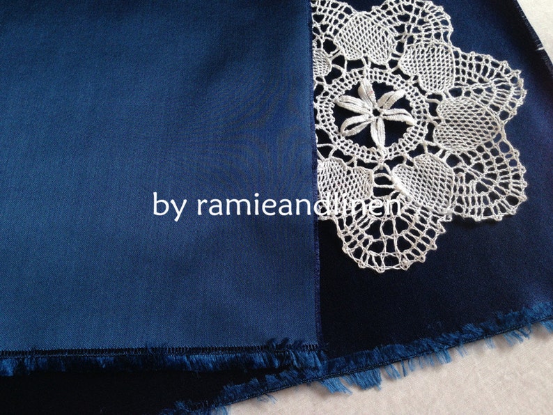 May include: A close-up of a dark blue fabric with a white crocheted lace doily on top. The fabric has a fringed edge.