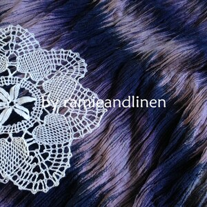 Puede incluir: Doily de ganchillo blanco con un delicado dise&ntilde;o floral, descansando sobre una tela de color p&uacute;rpura y azul tie-dye.