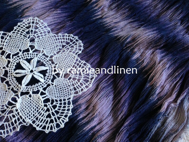 Puede incluir: Doily de ganchillo blanco con un delicado dise&ntilde;o floral, descansando sobre una tela de color p&uacute;rpura y azul tie-dye.