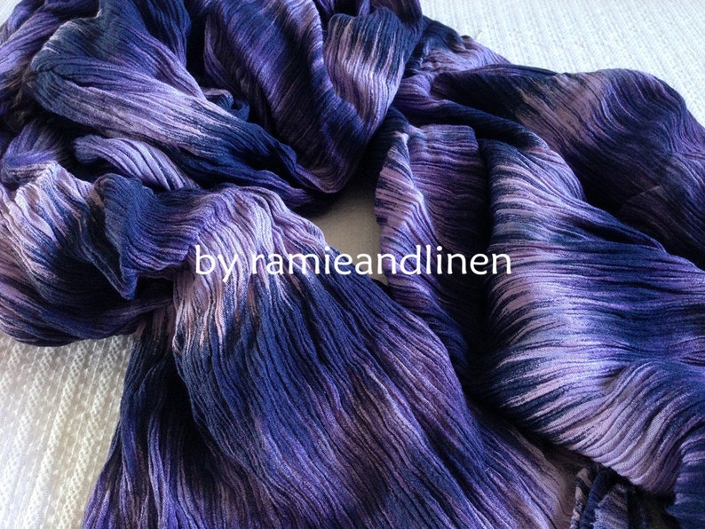 Puede incluir: Una bufanda de rayas moradas y azules con textura arrugada. La bufanda est&aacute; doblada por la mitad y el texto "by hamieandlinen" es visible en el centro.