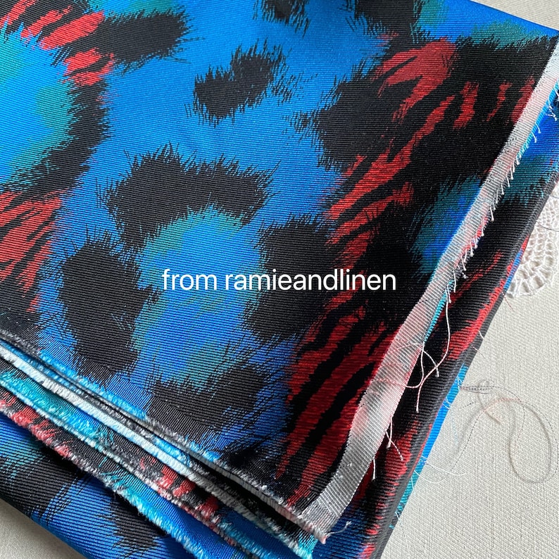May include: Close-up of a folded fabric with a blue, black, red, and green abstract print. The fabric has a textured surface and is folded in thirds. The text "from ramieandlinen" is visible in the image.