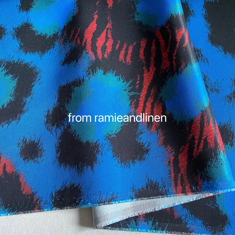 May include: Blue fabric with a black, red, and green abstract print. The fabric has a textured surface and is folded in half. The text "from ramieandlinen" is printed on the fabric.