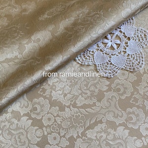 May include: A close-up of a beige fabric with a white floral damask pattern. The fabric is folded over itself and a white crocheted doily is on top of the fabric. The text "from ramieandlinen" is visible in the lower left corner of the image.