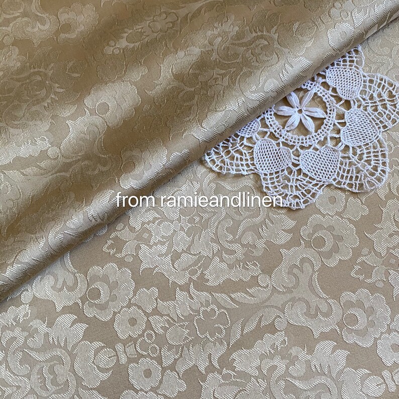 May include: A close-up of a beige fabric with a white floral damask pattern. The fabric is folded over itself and a white crocheted doily is on top of the fabric. The text "from ramieandlinen" is visible in the lower left corner of the image.