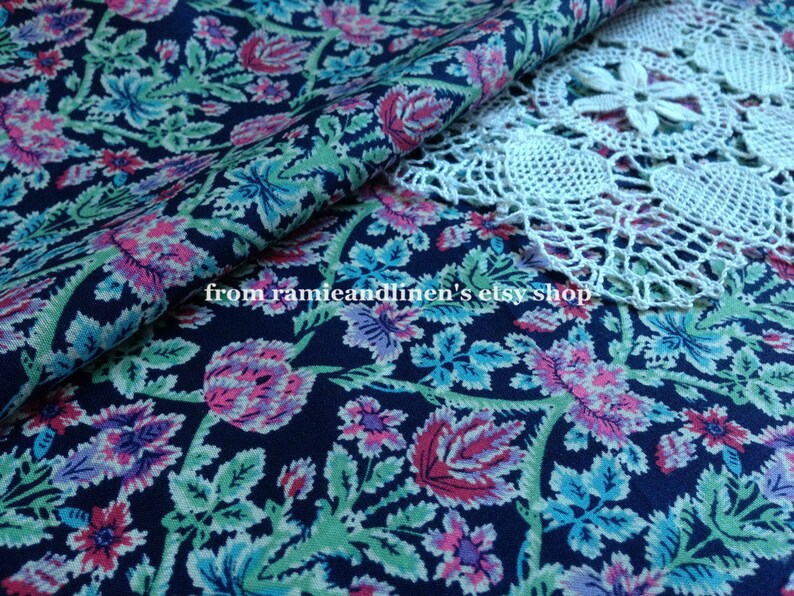 May include: A close-up of a dark blue fabric with a floral pattern. The fabric features pink, blue, and green flowers on a dark blue background. The fabric is folded in half and a white crocheted doily is partially visible in the top right corner. The text "from ramieandlinen's etsy shop" is visible in the lower left corner.