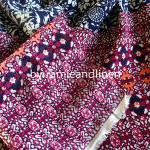 Pu&ograve; includere: Primo piano di un tessuto colorato con un motivo ripetitivo di piccoli fiori bianchi e blu su uno sfondo blu scuro. Il tessuto &egrave; piegato, rivelando un secondo strato con un motivo rosa e arancione di piccoli fiori bianchi su uno sfondo rosso scuro. Il testo "by ramieandlinen" &egrave; visibile al centro dell'immagine.
