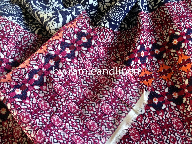 Pu&ograve; includere: Primo piano di un tessuto colorato con un motivo ripetitivo di piccoli fiori bianchi e blu su uno sfondo blu scuro. Il tessuto &egrave; piegato, rivelando un secondo strato con un motivo rosa e arancione di piccoli fiori bianchi su uno sfondo rosso scuro. Il testo "by ramieandlinen" &egrave; visibile al centro dell'immagine.