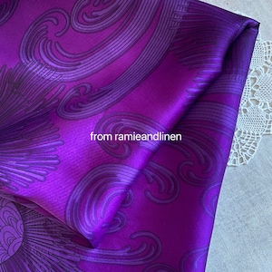 Può includere: Tessuto di raso viola con un motivo a spirale astratto. Il tessuto è piegato a metà e il testo "from ramieandlinen" è visibile.