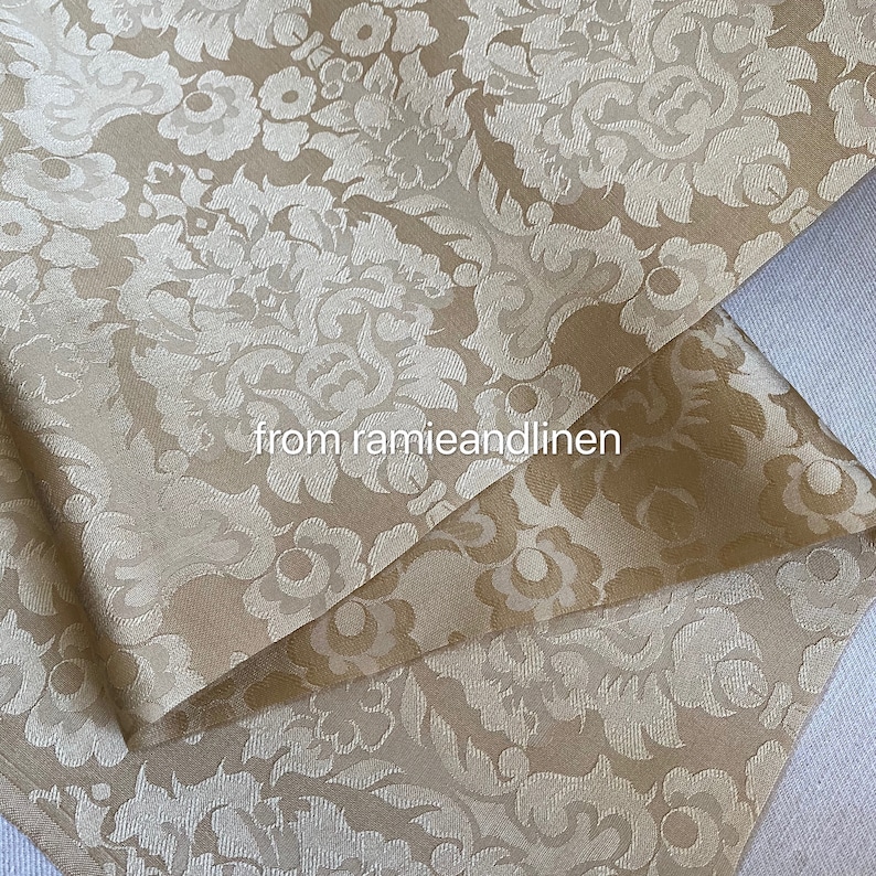 May include: A close-up of a beige fabric with a white damask pattern. The fabric is folded in half, showing the pattern on both sides. The text "from ramieandlinen" is visible in the center of the image.