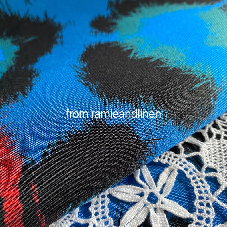 May include: Close-up of a blue fabric with a black, red, and green abstract print. The fabric has a textured weave and is folded over a white lace fabric.