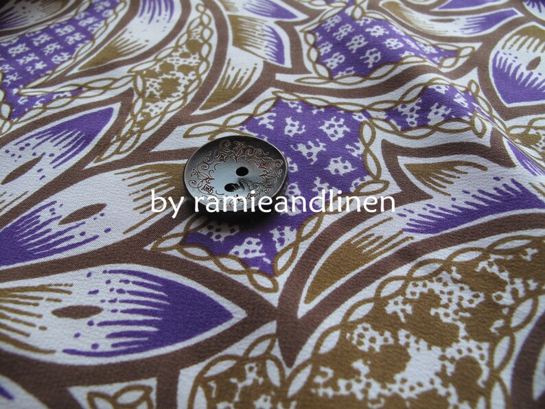 May include: A close-up of a brown and purple fabric with a white button in the center. The fabric has a repeating pattern of paisley-like shapes. The button is silver and has a decorative design.