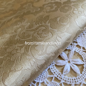 May include: A close-up of a gold damask fabric with a subtle sheen. The fabric is folded over a white crocheted lace doily. The text "from ramieandlinen" is visible in the image.