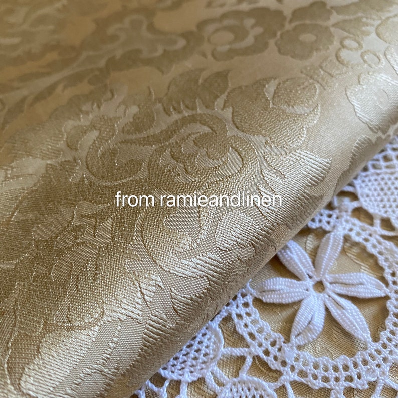 May include: A close-up of a gold damask fabric with a subtle sheen. The fabric is folded over a white crocheted lace doily. The text "from ramieandlinen" is visible in the image.