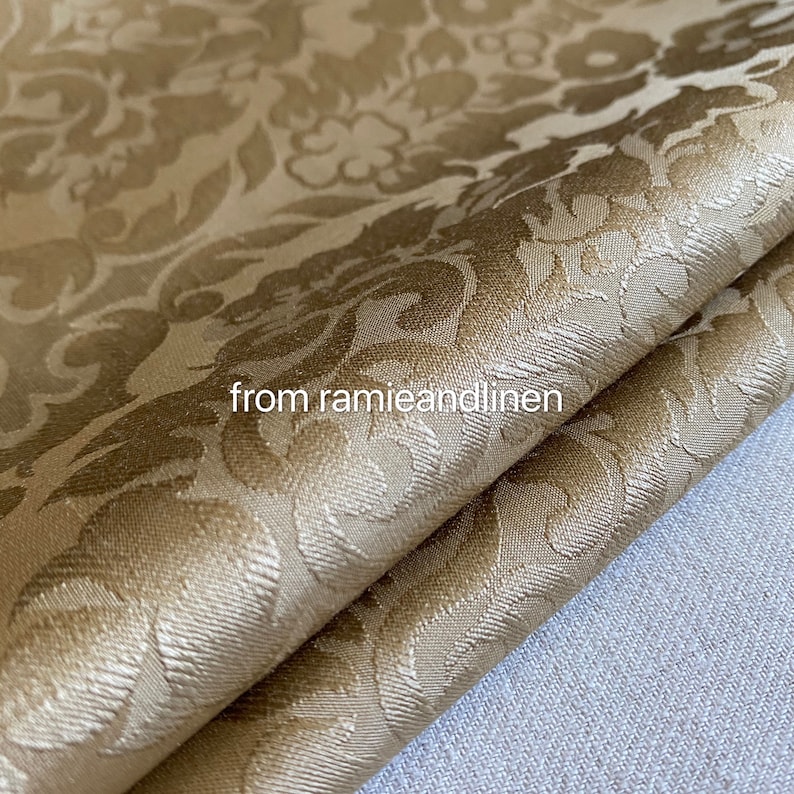 May include: A close-up of a gold fabric with a damask pattern. The fabric is folded in half and has a soft, luxurious feel.