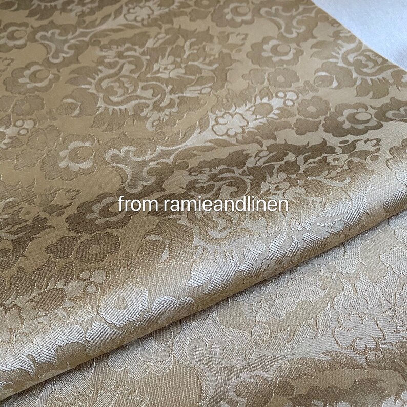 May include: A close-up of a beige fabric with a white damask pattern. The fabric is folded in half and has a subtle sheen.