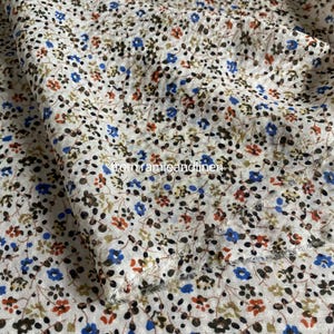 May include: Close-up of a white fabric with a floral pattern. The fabric is covered in small blue, red, and brown flowers with green leaves. The text "from ramieandlinen" is visible on the fabric.
