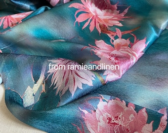 May include: A teal blue silk scarf with a pink floral print. The flowers are stylized and resemble cactus blossoms. The scarf is folded and draped over a white surface. The text "from ramieandlinen" is printed on the scarf.