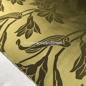 Op de afbeelding: Een close-up van een gouden stof met een donkerbruin bloemenpatroon. De stof heeft een subtiele glans en een zachte textuur.