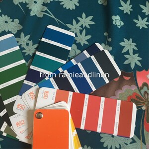 K&ouml;nnte beinhalten: Eine Nahaufnahme eines Stoffmusters mit einem floralen Muster in den Farben Teal, Gr&uuml;n und Wei&szlig;. Ein F&auml;cher mit Farbmustern liegt auf dem Stoff. Die Farbmuster sind mit RAL-Nummern und Farbnamen beschriftet, wie z. B. "Copblau", "Ceanblau", "Smarag", "Azublau", "Endbeenot", "Rond" und "Korolierect".