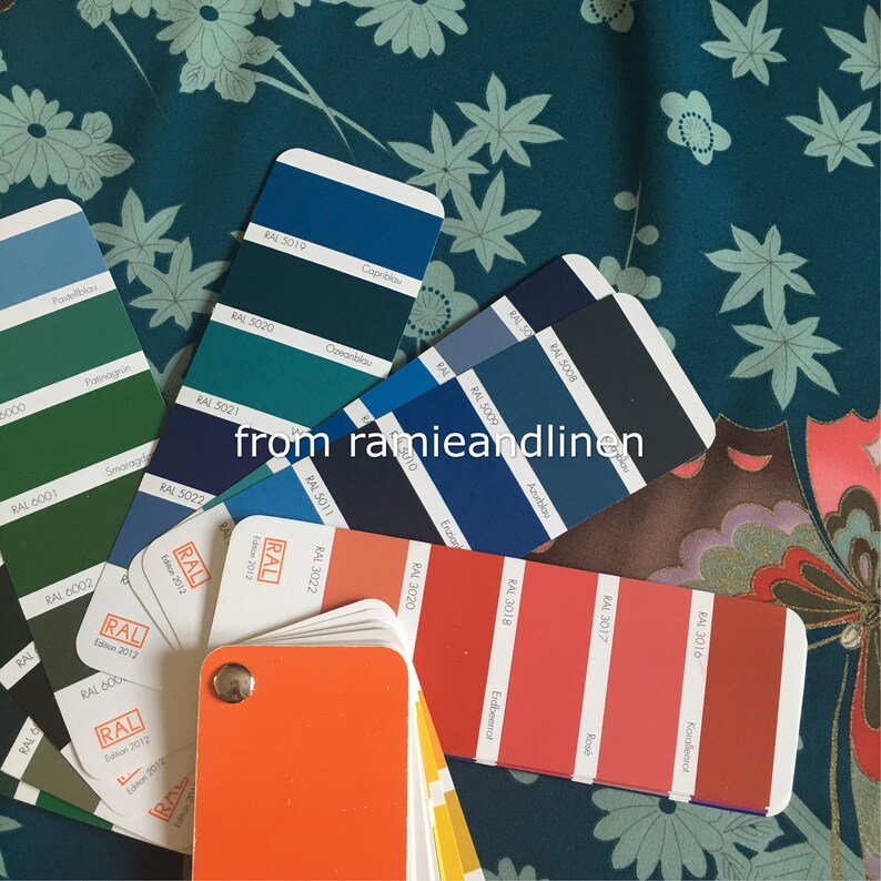 K&ouml;nnte beinhalten: Eine Nahaufnahme eines Stoffmusters mit einem floralen Muster in den Farben Teal, Gr&uuml;n und Wei&szlig;. Ein F&auml;cher mit Farbmustern liegt auf dem Stoff. Die Farbmuster sind mit RAL-Nummern und Farbnamen beschriftet, wie z. B. "Copblau", "Ceanblau", "Smarag", "Azublau", "Endbeenot", "Rond" und "Korolierect".