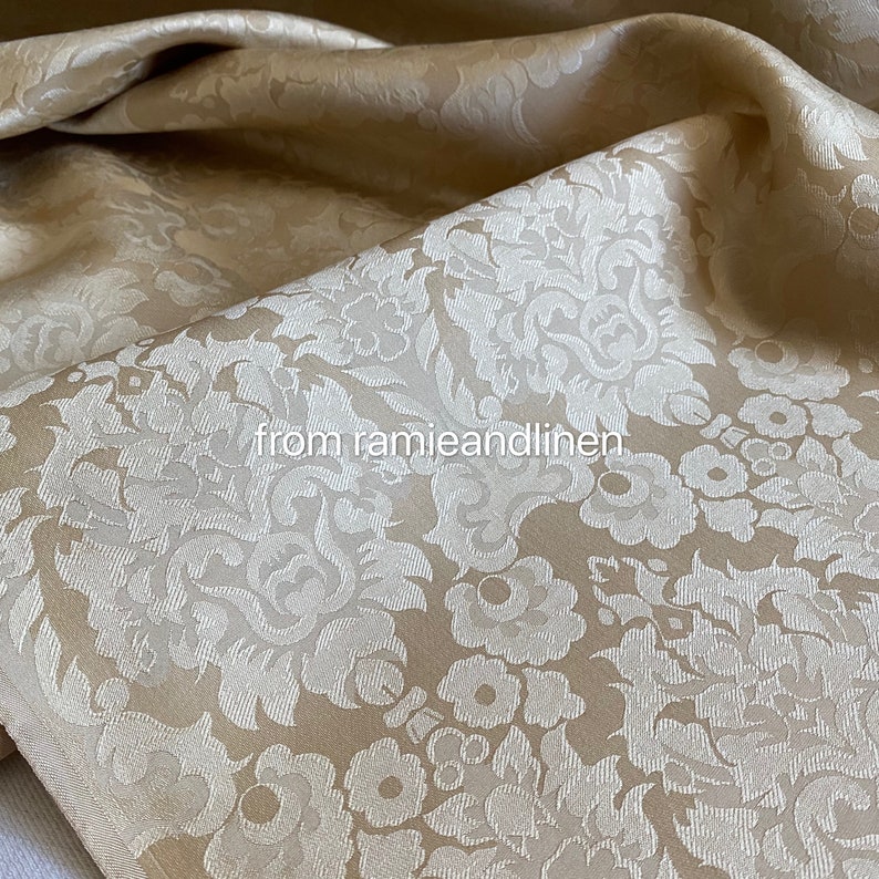 May include: A close-up of a beige fabric with a white damask pattern. The fabric is soft and luxurious, and it would be perfect for upholstery or curtains.