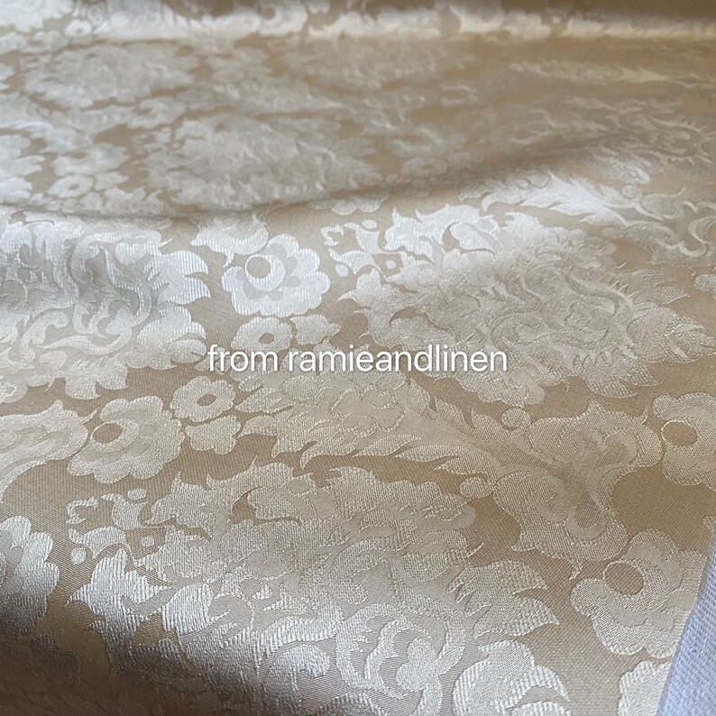 May include: A close-up of a beige fabric with a white damask pattern. The fabric has a subtle sheen and a soft texture. "from ramieandlinen" is printed on the fabric.