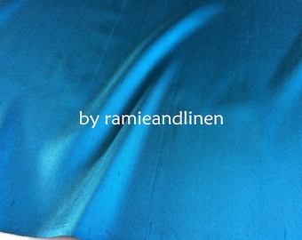 May include: A close-up of a smooth, teal blue fabric with a subtle sheen. The fabric is draped and folded, creating soft curves. The text "by ramieandlinen" is printed in white in the center of the image.