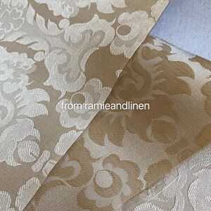 May include: Close-up of a beige fabric with a white damask pattern. The fabric is folded in half, showing the pattern on both sides. The fabric is from "ramieandlinen".