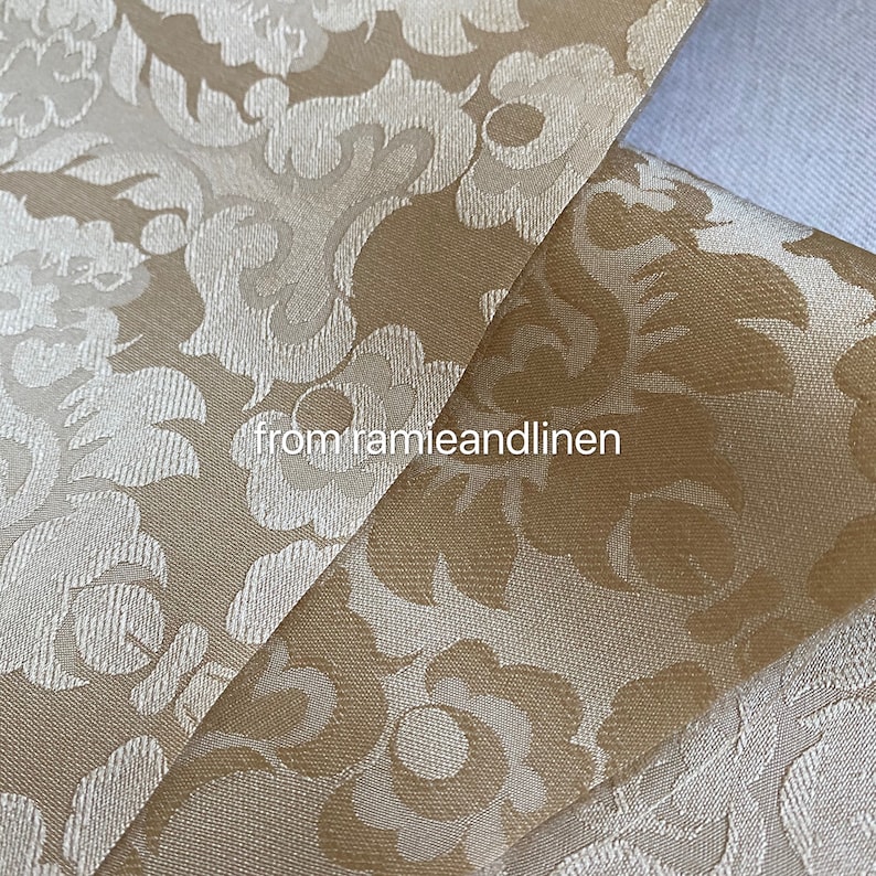 May include: Close-up of a beige fabric with a white damask pattern. The fabric is folded in half, showing the pattern on both sides. The fabric is from "ramieandlinen".