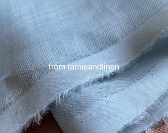 Puede incluir: Primer plano de una tela azul claro con una superficie ligeramente texturizada. La tela está doblada y los bordes están deshilachados. El texto "from ramieandlinen" es visible en la imagen.