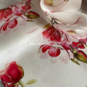 Puede incluir: Tela de seda blanca con estampado floral de flores de magnolia rosas y rojas. La tela es suave y fluida, y las flores son delicadas y realistas.