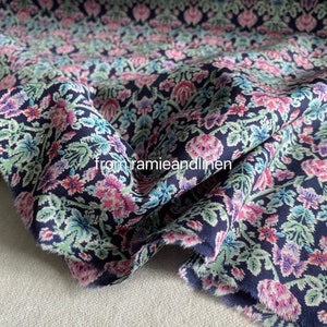 May include: A close-up of a navy blue fabric with a floral print. The fabric features a repeating pattern of pink, white, and blue flowers and leaves. The fabric is soft and has a slightly textured surface.