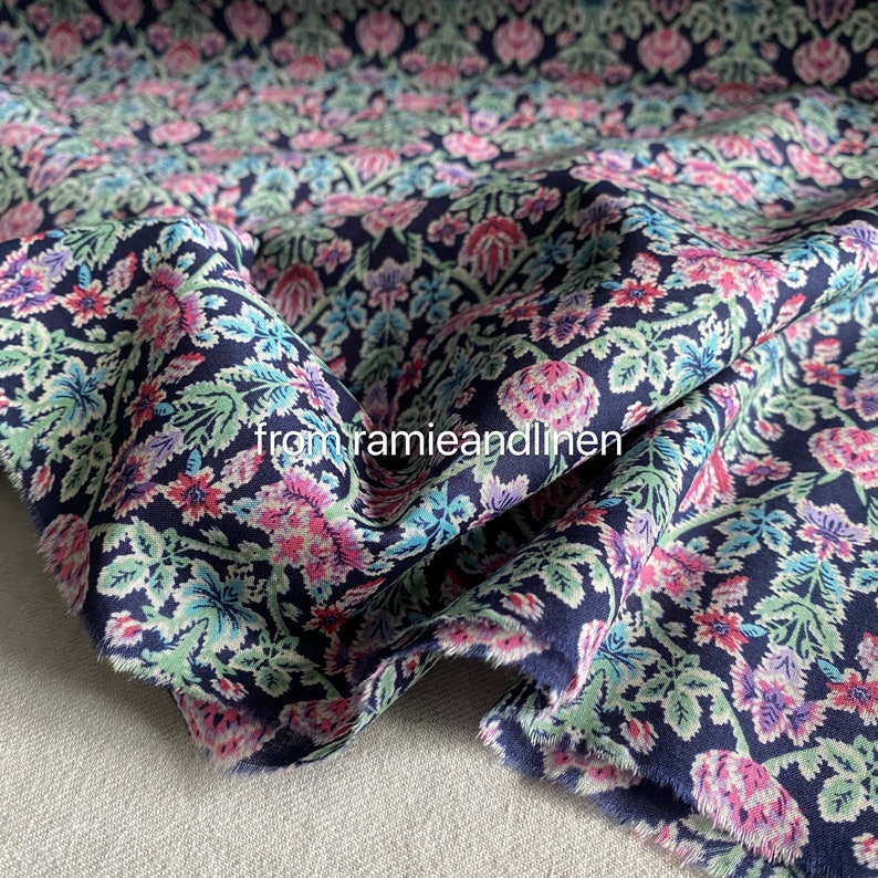 May include: A close-up of a navy blue fabric with a floral print. The fabric features a repeating pattern of pink, white, and blue flowers and leaves. The fabric is soft and has a slightly textured surface.