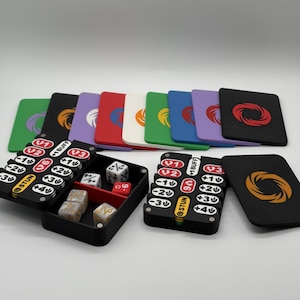 Può includere: Una collezione di accessori per il gioco, tra cui dadi, porta carte e carte da gioco. Le carte sono nere con un design circolare in vari colori. I dadi sono bianchi e beige. I porta carte sono neri con testo rosso, bianco e giallo.