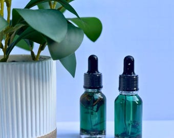 May include: Two small glass dropper bottles filled with a green liquid, set before a white and tan ceramic pot with a green plant. The bottles have black droppers and contain a clear liquid with green herbs.