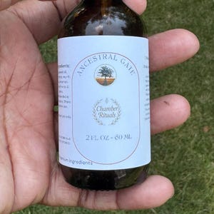 May include: A brown glass bottle with a black cap, held in a hand. The label reads "ANCESTRAL GATE" and "Chamber Rituals." The bottle contains 2 fl oz, equivalent to 60 ml. The background is blurred green.