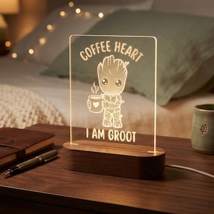 Può includere: Una luce notturna a LED rettangolare in acrilico con base in legno, caratterizzata dall'incisione di un piccolo personaggio simile a un albero che tiene una tazza di caffè. Il testo COFFEE HEART e I AM GROOT è inciso sul pannello trasparente.