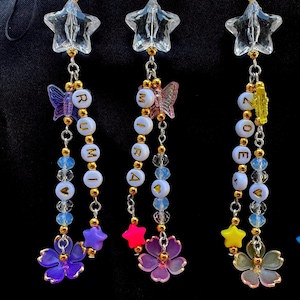 May include: Three beaded star charms with the word "RUMI" and "ZOE" spelled out in gold letters. Each charm features a clear star, butterfly, flower, and heart-shaped beads in various colors, suspended by gold chains.