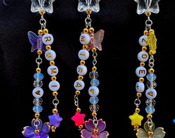 May include: Three beaded star charms with the word "RUMI" and "ZOE" spelled out in gold letters. Each charm features a clear star, butterfly, flower, and heart-shaped beads in various colors, suspended by gold chains.
