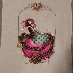 May include: Cross-stitch embroidery of a woman with long brown hair sitting in a pink and brown swirl. The woman is wearing a green dress with a floral pattern and a crown of flowers. The embroidery is framed by a floral arch with the letter "R" in the center.