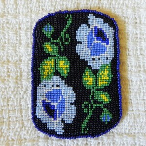 May include: A close-up of a beaded floral design with blue, white, yellow, and green beads on a black background. The design is surrounded by a dark blue border.