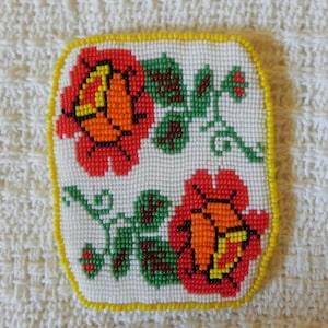 May include: A square, beaded coaster with a white background and a floral design. The flowers are red and orange, with green leaves and stems. The border is yellow.