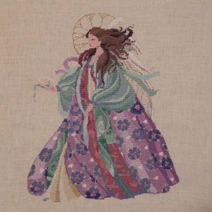 May include: A cross-stitch embroidery of an angel with a halo, wearing a flowing gown with purple and green floral patterns. The angel is holding a small bird in its hand.