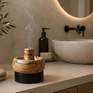 Puede incluir: Un quemador de incienso de madera de color marrón claro con vetas naturales oscuras y una chimenea central que emite un fino hilo de humo. El quemador se asienta sobre una base cilíndrica negra sólida, creando un contraste con la madera natural.