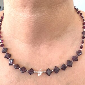 May include: A necklace featuring dark red, square-shaped beads with carved floral designs. The beads are strung together with small silver-colored beads. A clear, irregular-shaped bead is also included in the necklace's design.