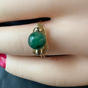 May include: A gold-toned ring featuring a large, round, deep green bead as its focal point. The bead is securely held within a wire-wrapped setting. The ring's design is simple yet elegant, showcasing the vibrant color of the gemstone.