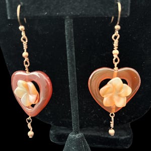 May include: A pair of heart-shaped earrings. Each earring features a translucent, reddish-brown heart with a flower in the center. The earrings are suspended from gold-colored hooks and have decorative wire and bead accents.