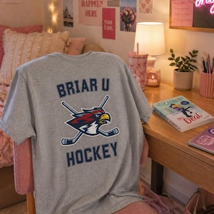 Op de afbeelding: Een heather grijze t-shirt met de tekst "BRIAR U HOCKEY" en een afbeelding van een havik en gekruiste hockeysticks. Het shirt wordt getoond op een roze stoel in een kamer met een bureau en decoratieve items.