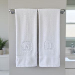 Può includere: Due asciugamani da bagno rettangolari bianchi appesi a un portasciugamani in metallo cromato. Ogni asciugamano presenta un bordo testurizzato sul fondo e un monogramma ricamato centrato in una cornice quadrata, con la lettera M e la lettera H.