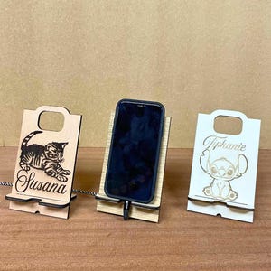 Puede incluir: Tres soportes de teléfono de madera. Uno presenta una ilustración de gato y el nombre "Susana". Otro sostiene un smartphone negro. El tercer soporte tiene un personaje de dibujos animados y el nombre "Tiphanie". Todos los soportes tienen un asa.